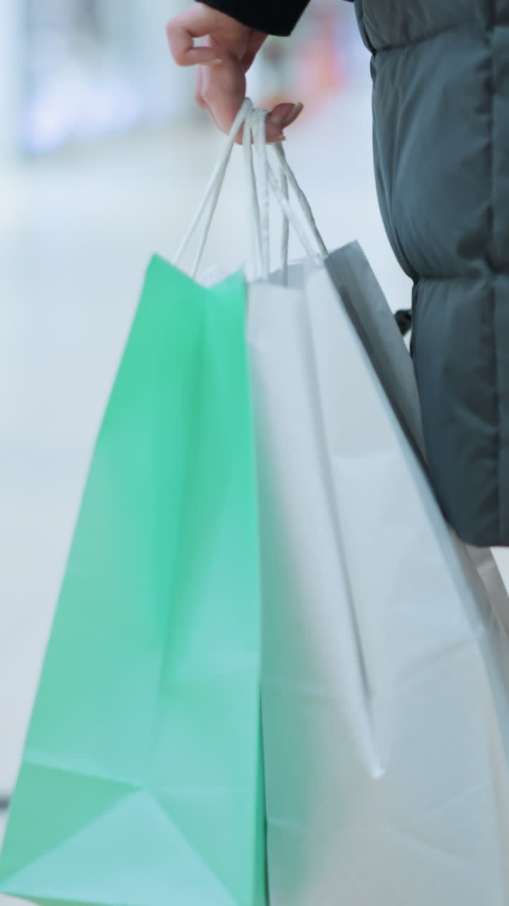 vista trasera de una persona con pantalones vaqueros y chaqueta negra caminando en un espacio comercial bien iluminado llevando un bolso de mano negro y dos bolsas de compras, fondo borroso con pantallas de venta al por menor brillantes