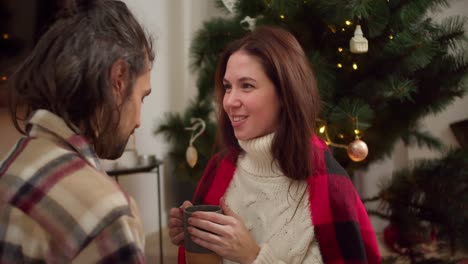 Close-up-shot-of-a-guy-and-a-brunette-girl-in-a-checkered-shirt-and-a-red-blanket-sitting-talking-and-looking-at-each-other,-a-girl-drinking-a-hot-drink-from-a-gray-glass-near-a-green-decorated-Christmas-tree-in-the-winter-evening