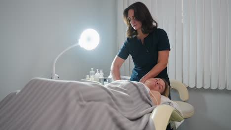 wide view of skin care expert gliding fingertips along side of patient neck toward shoulder during relaxing spa session inside bright clinic room with subtle soft product trolley in background