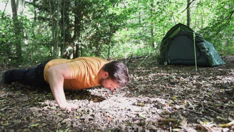 young strong man doing push ups in forest campsite with tent in background