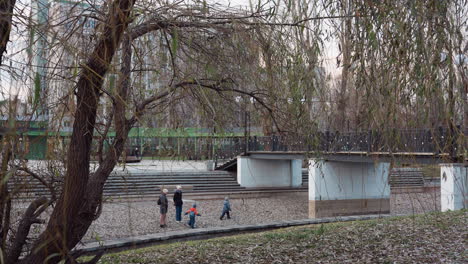 leafy winter tree branches hang in foreground above stone path as people walk near pedestrian bridge in quiet urban park, soft evening light and distant buildings evoke peaceful seasonal atmosphere