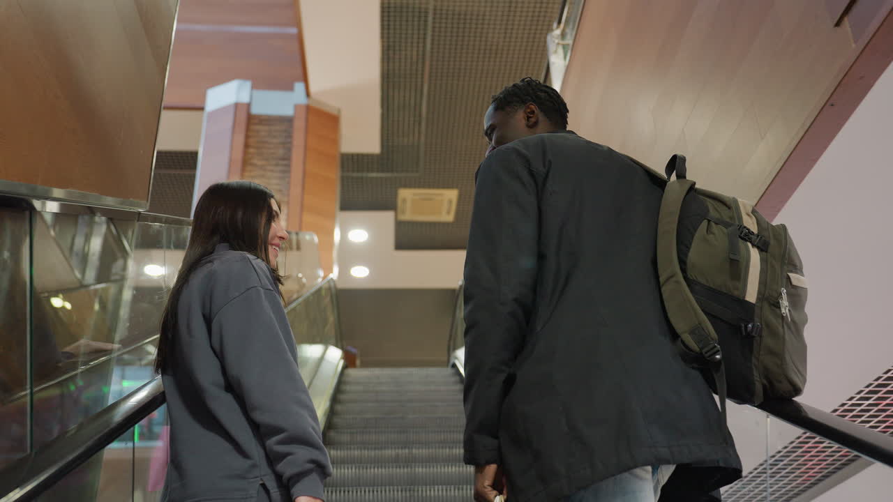 Two business colleagues riding up escalator inside modern building, engaging in cheerful conversation, wearing casual jackets and carrying shopping bags and backpack