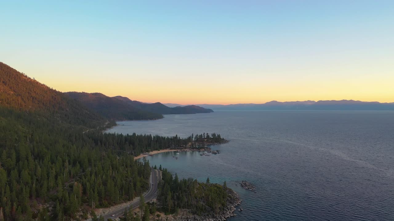 Colorful Sunset Over Lake Taho with a Road or Highway Going Through a Pine Tree Forest with Clear Blue Water and Mountains in the Background - aerial drone view