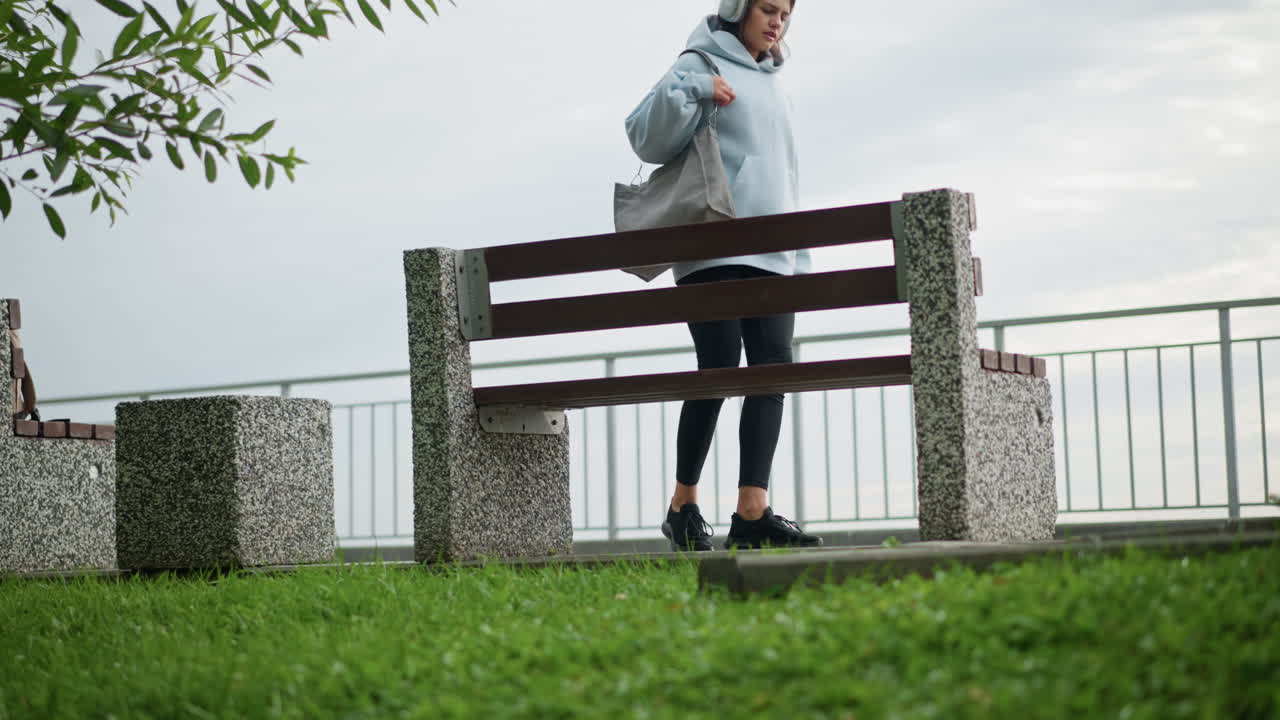 Pretty woman in casual wear walking gracefully toward concrete bench, sitting down and crossing her legs with bag beside her in serene outdoor environment, surrounded by grass and leaves