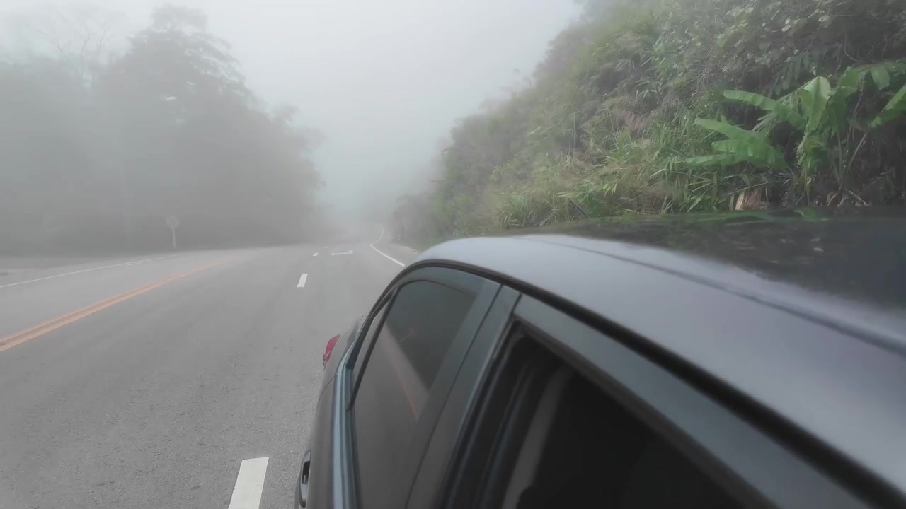 Ripresa in soggettiva guidando lungo un'autostrada asfaltata bagnata e tortuosa, avvolta in una fitta nebbia mattutina a Phetchabun. Caratterizzata da lussureggianti alberi tropicali verdi e cartelli stradali in montagna.