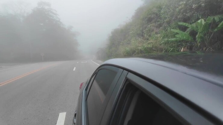 Ripresa in soggettiva guidando lungo un'autostrada asfaltata bagnata e tortuosa, avvolta in una fitta nebbia mattutina a Phetchabun. Caratterizzata da lussureggianti alberi tropicali verdi e cartelli stradali in montagna.
