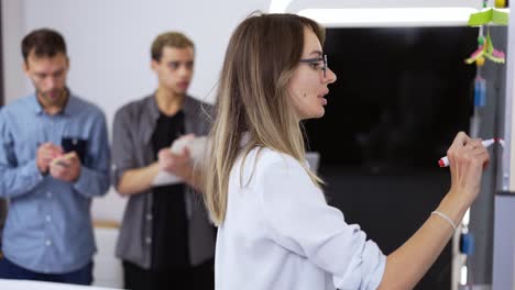 una joven trabajadora de oficina de pie y escribiendo en una pizarra blanca hablando con sus colegas