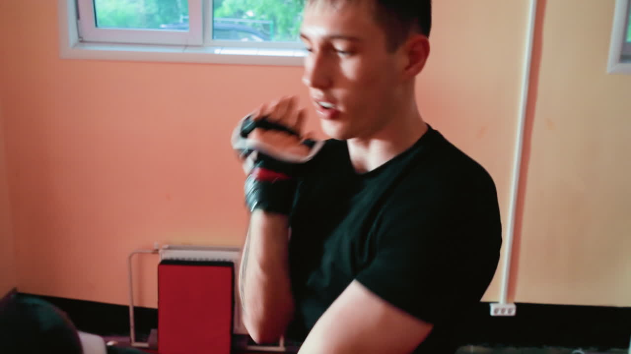 Karate practitioners engaged in sparring session inside dojo, focused athlete in black shirt wearing gloves holding strong guard stance while opponent approaches, showcasing discipline, concentration
