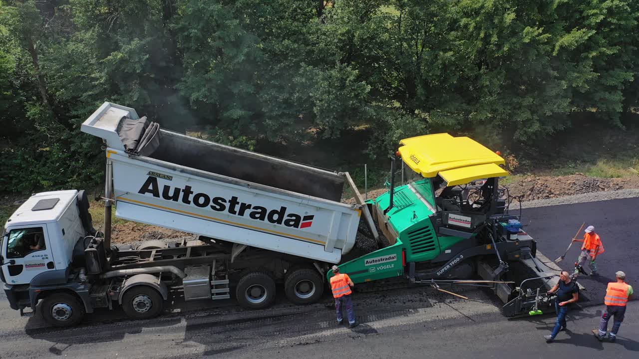 Road works on asphalt pavement construction. Heavy machinery on the road. Workers in orange uniform leveling up hot asphalt on the highway. Aerial view.