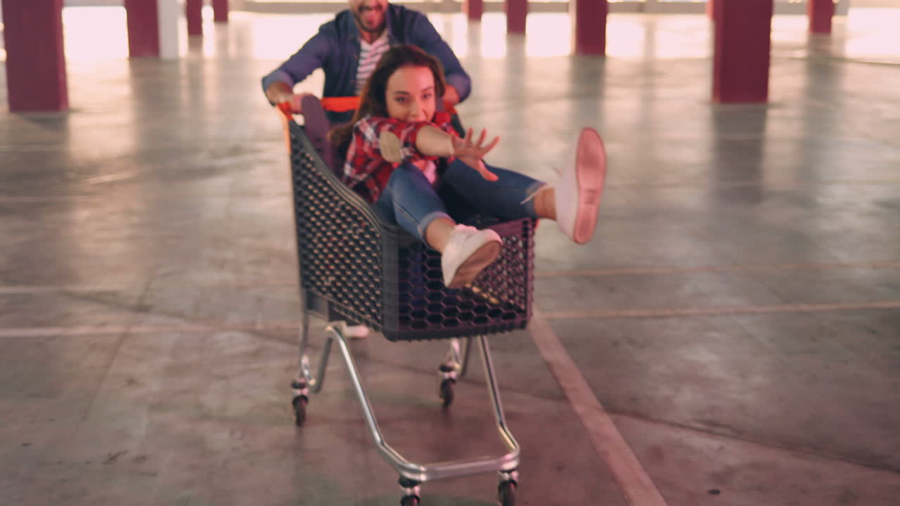 젊고 행복한 백인 소녀들이 트롤리에 앉아 웃고 빈 주차장에서 남자친구에게 실려 가는 모습을 가까이서 볼 수 있습니다.