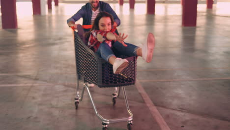 close-up view of young and happy caucasian girls sitting in trolleys, laughing and being carried by their boyfriends in the empty parking