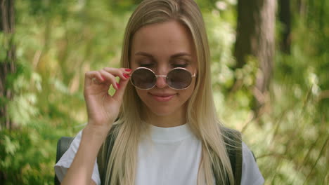 a young female traveler in sunglasses looks directly at the camera and smiles flirting a traveler with a backpack in the park and in the forest in slow motion