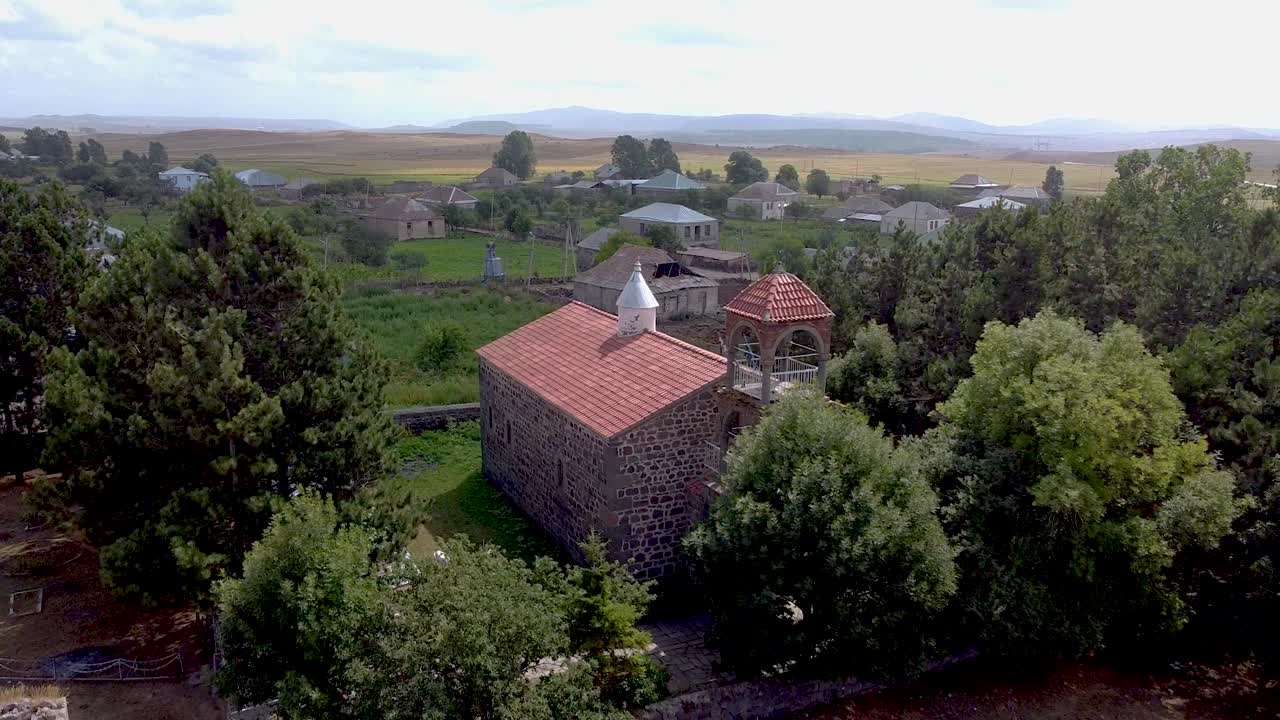 A beautiful stone church sits in a green landscape, its red roof contrasting with the trees. Nearby houses and mountains complete this tranquil rural scene