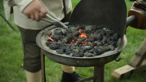 a revolutionary war reinacter blacksmith puts a piece of metal in hot coals to heat up while he pumps the bellows