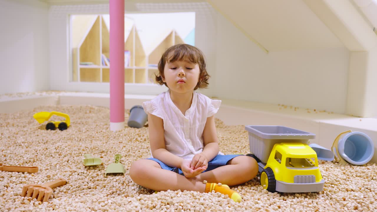 Mixed Korean girl playing with a toy truck in a modern indoor sandbox with wooden pellets at a play center - slow motion