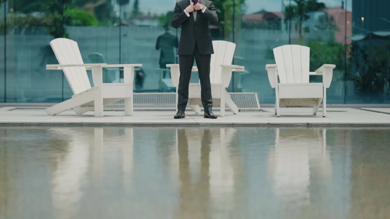 Groom in elegant suit adjusting jacket near poolside with reflection in water