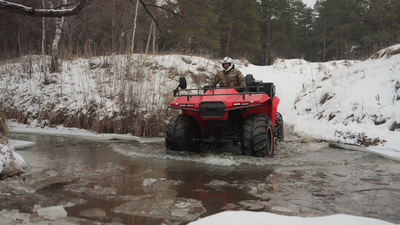 ATV rosso che attraversa un torrente ghiacciato in una foresta innevata, pilota con casco naviga in acque ghiacciate poco profonde, pneumatici grandi che smuovono fango, acceleratore cauto, neve che cade, brivido teso e controllato, avventuriero solitario.