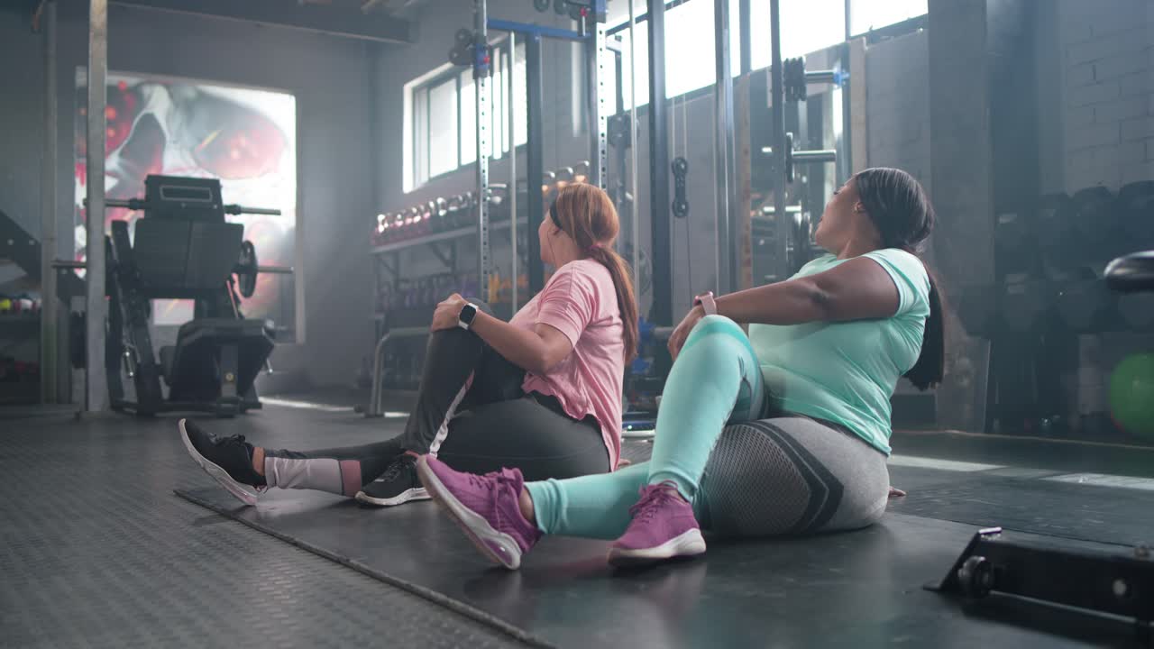 Diverse women smiling and talking, performing seated torso twists on black mat at gym warming up