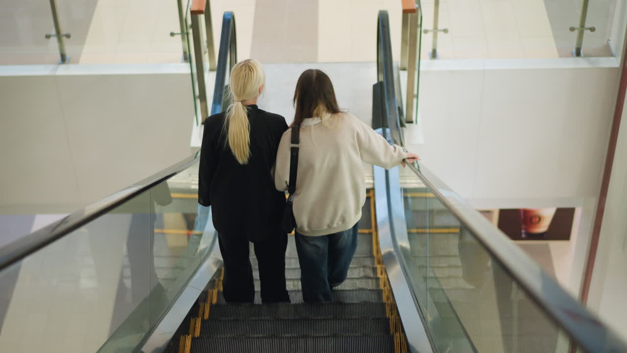 Back view of two sisters descending escalator inside modern shopping mall, walking closely together while holding onto railing, creating warm candid scene of companionship