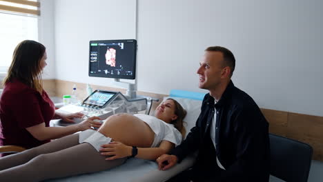 pregnant woman lying on doctor's couch is happy to see her unborn baby on the monitor. caucasian couple expecting a child.