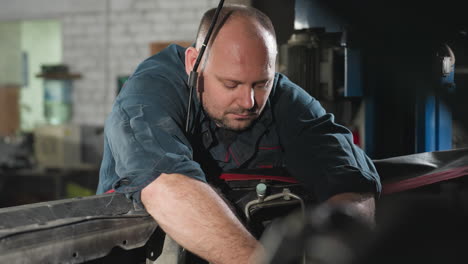 engineer in blue uniform works on car engine in mechanical workshop, focused on repair task, hands-on automotive maintenance in industrial setting with tools and machinery in background