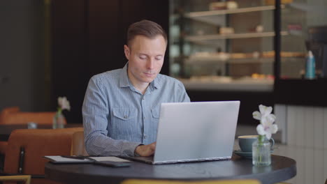 Un-Joven-Con-Camisa-Está-Sentado-En-Una-Mesa-Con-Una-Computadora-Portátil-Y-Escribiendo-En-El-Teclado.-Un-Estudiante-Puede-Estudiar-De-Forma-Remota.-Un-Empresario-Realiza-Su-Negocio-De-Forma-Remota