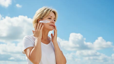 a woman applying a facial cream outdoors under a bright blue sky with fluffy clouds, showcasing a serene moment dedicated to skincare and self-care.