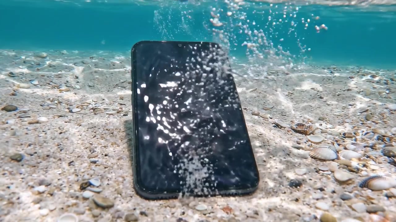 An underwater perspective of a smartphone resting on the sandy seabed, surrounded by pebbles and bubbles while light refracts in the clear blue water above.
