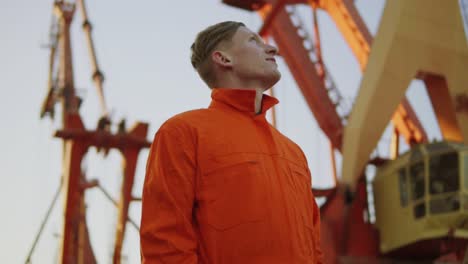 Hübscher-Junger-Containerlagerarbeiter-In-Orangefarbener-Uniform,-Der-Neben-Dem-Schiff-Im-Hafen-Steht-Und-Nach-Oben-Schaut.-Großer-Kran-Am
