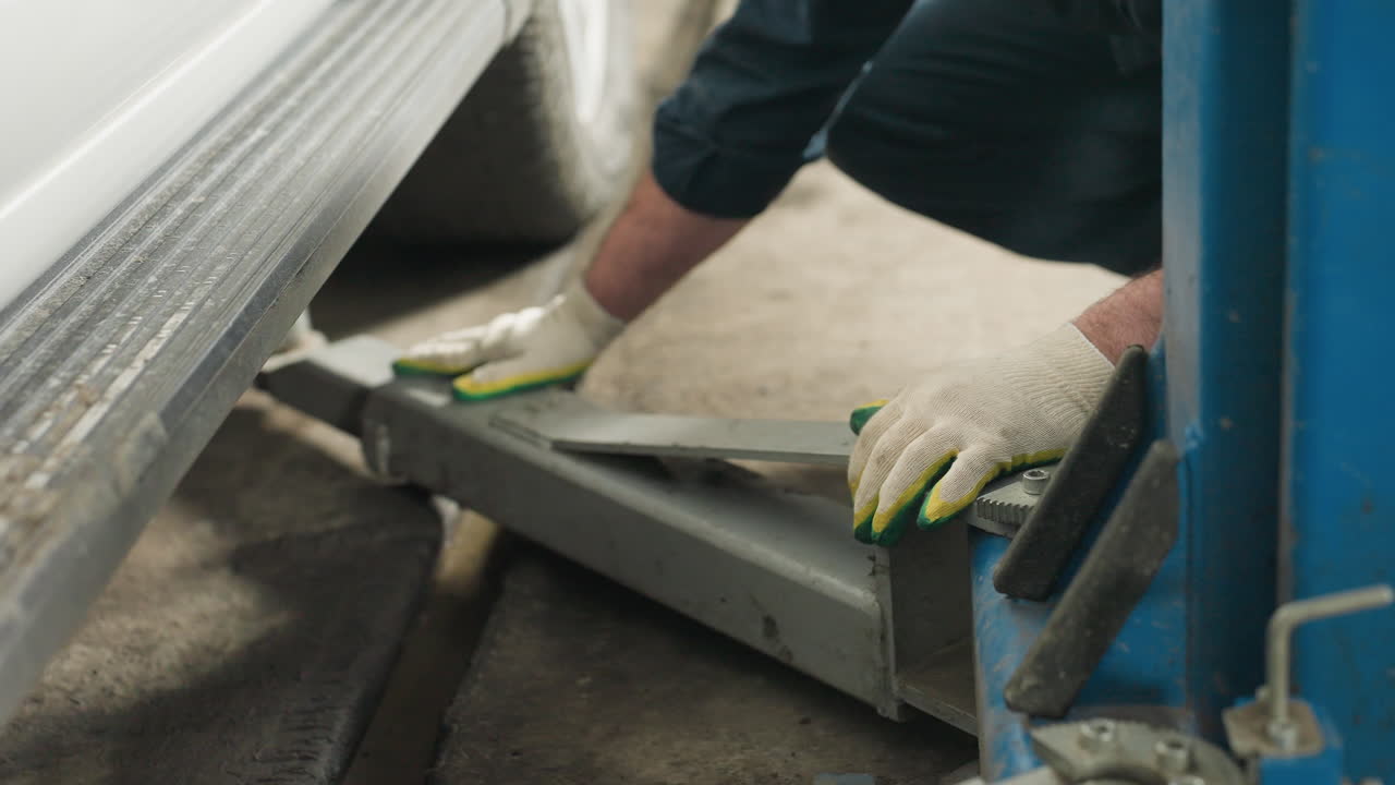 Close-up of a mechanic's hand in gloves positioning a car jack under a vehicle next to a blue lift in a well-equipped garage, preparing for maintenance work