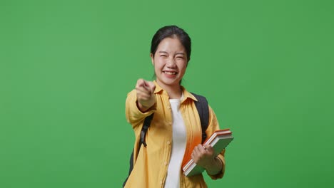 asian woman student with a backpack and some books touching her chest then smiling and pointing to camera being proud of herself in the green screen background studio