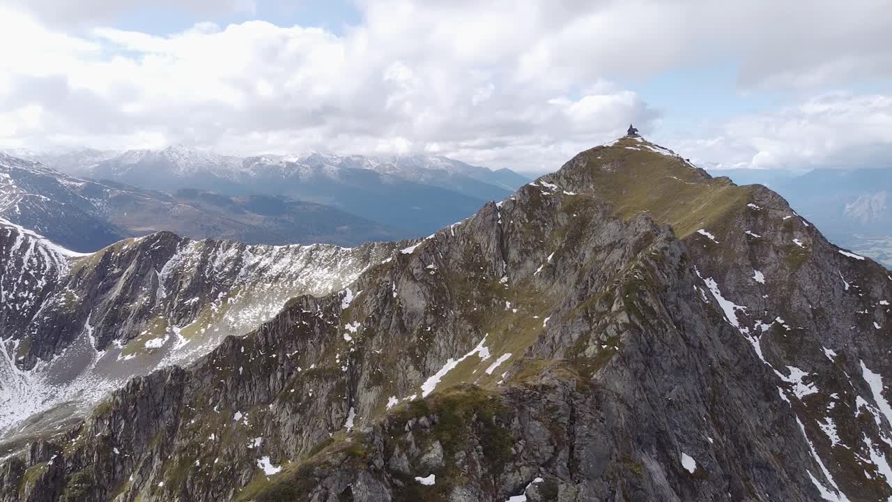 rápido vuelo aéreo alrededor de la cima de una montaña alpina rocosa y empinada con una vieja capilla de madera en el pico