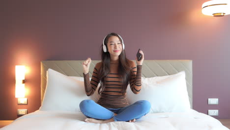 a pretty young woman sitting on the bed of her hotel suite dances to the music from her headphones