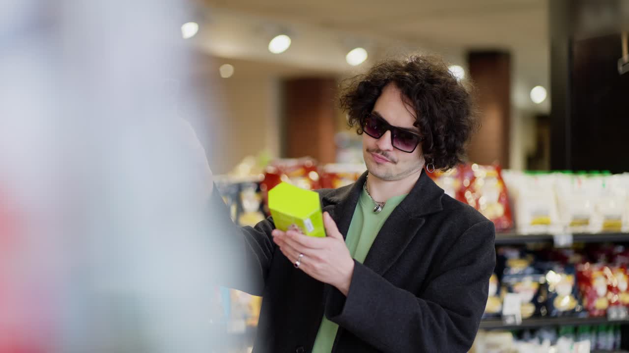A confident and cool guy with curly hair in black glasses and a black coat chooses the products he needs in a supermarket, choosing from two