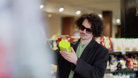 a confident and cool guy with curly hair in black glasses and a black coat chooses the products he needs in a supermarket, choosing from two