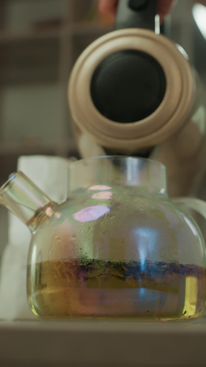 steaming glass teapot on counter, kettle pouring warm amber infusion with delicate condensation and soft bokeh herbal leaves settle in transparent vessel, cozy kitchen atmosphere, tea sommelier