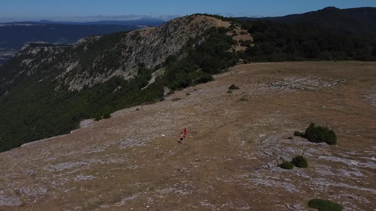 Two people followed by a drone while they run and practice trail running. One runner is without a shirt and the other runner is wearing a red shirt