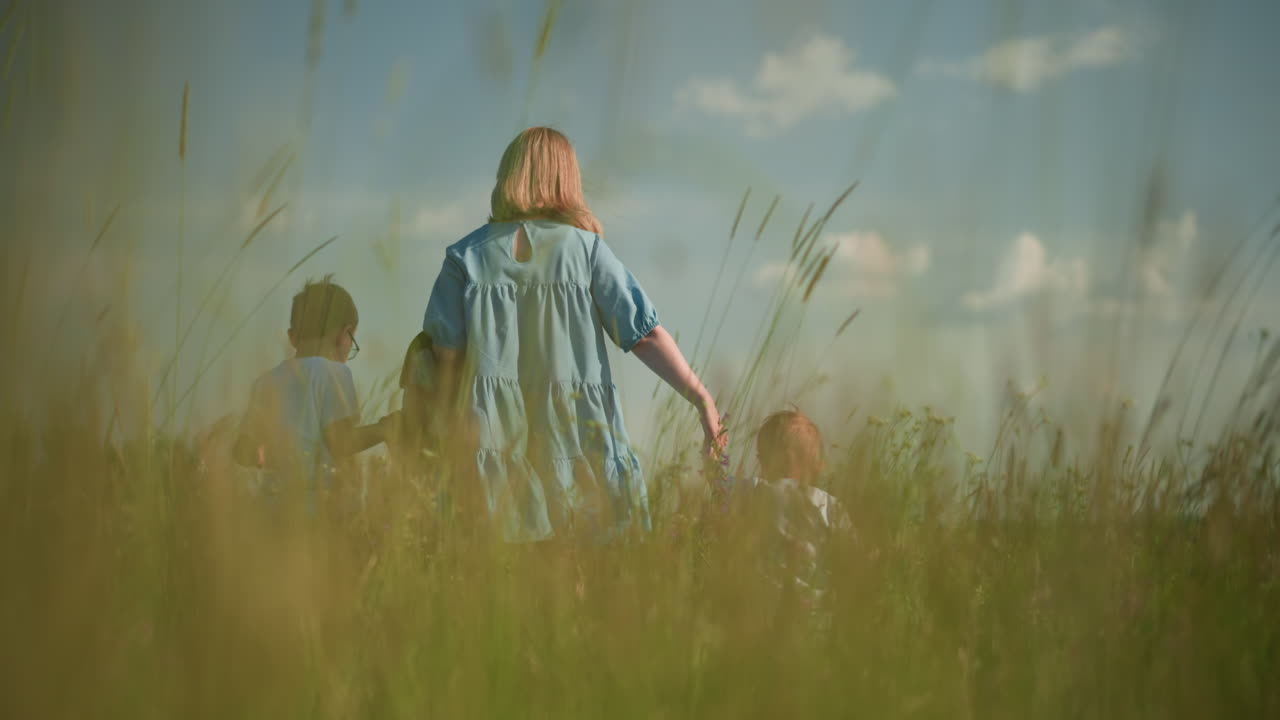 Back view of a woman in a flowing blue gown walking with her two young sons, both in white shirts, through a lush green grass field under a clear blue sky with scattered clouds