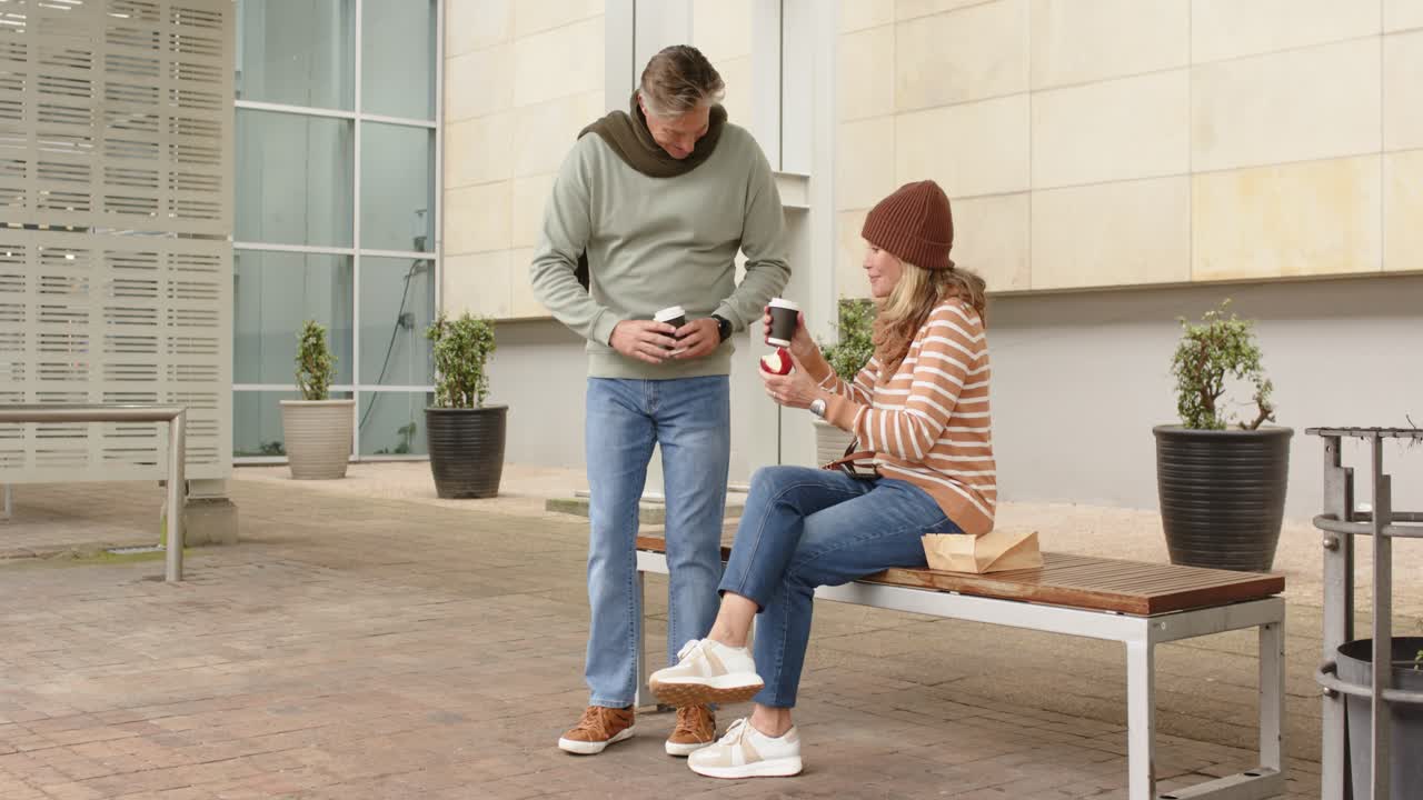 Pareja de mediana edad acercándose a un banco al aire libre; la mujer, con una manzana en la mano, recibe una taza de café y ambos comparten la bebida