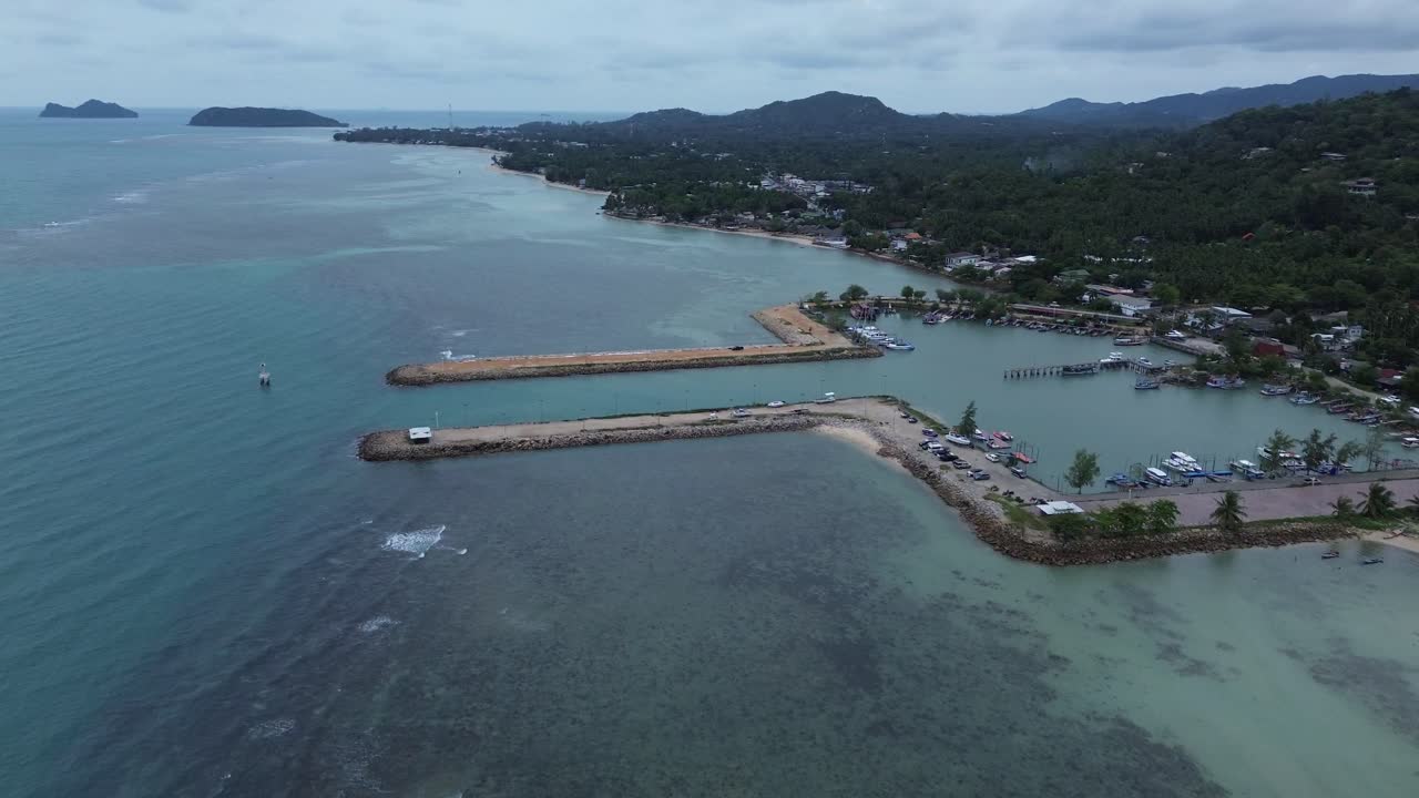Aerial drone footage showing Baan Tai Pier and the surrounding coastal town on Koh Phangan island, Surat Thani, Thailand, with tropical hills, beach shoreline, and blue sky