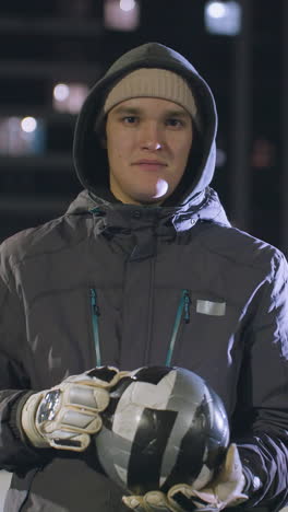 hombre con capucha y guantes golpeando casualmente una pelota de fútbol en su mano mientras está de pie en un campo deportivo urbano al aire libre, escena nocturna iluminada por luces bokeh y edificios de gran altura circundantes