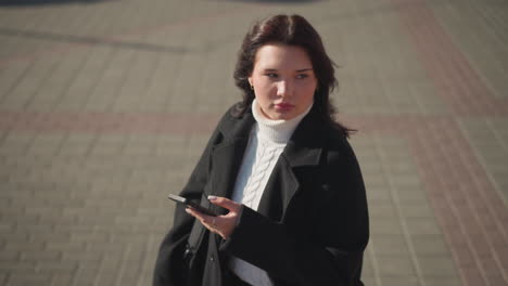 confident lady in white turtleneck and black coat focuses on her phone while walking on an interlocked path, occasionally looking around