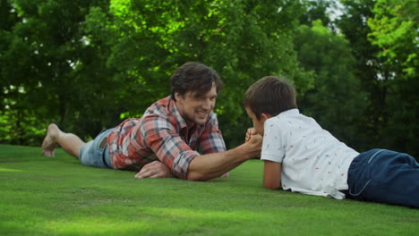 Father-and-son-lying-on-grass-in-field.-Man-and-boy-competing-in-arm-wrestling