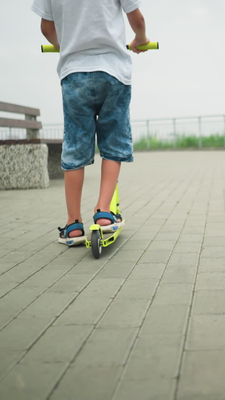 Back leg view of a woman and her children riding scooters outdoors on a paved path, with iron rail by the side