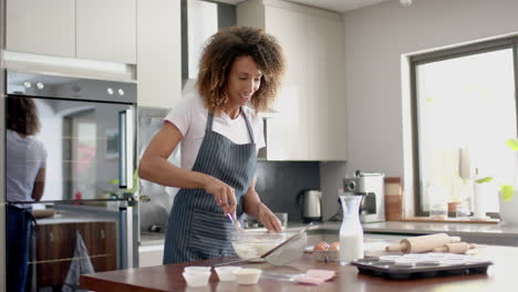 happy biracial woman in apron mixing cake mix, baking in kitchen, copy space, slow motion