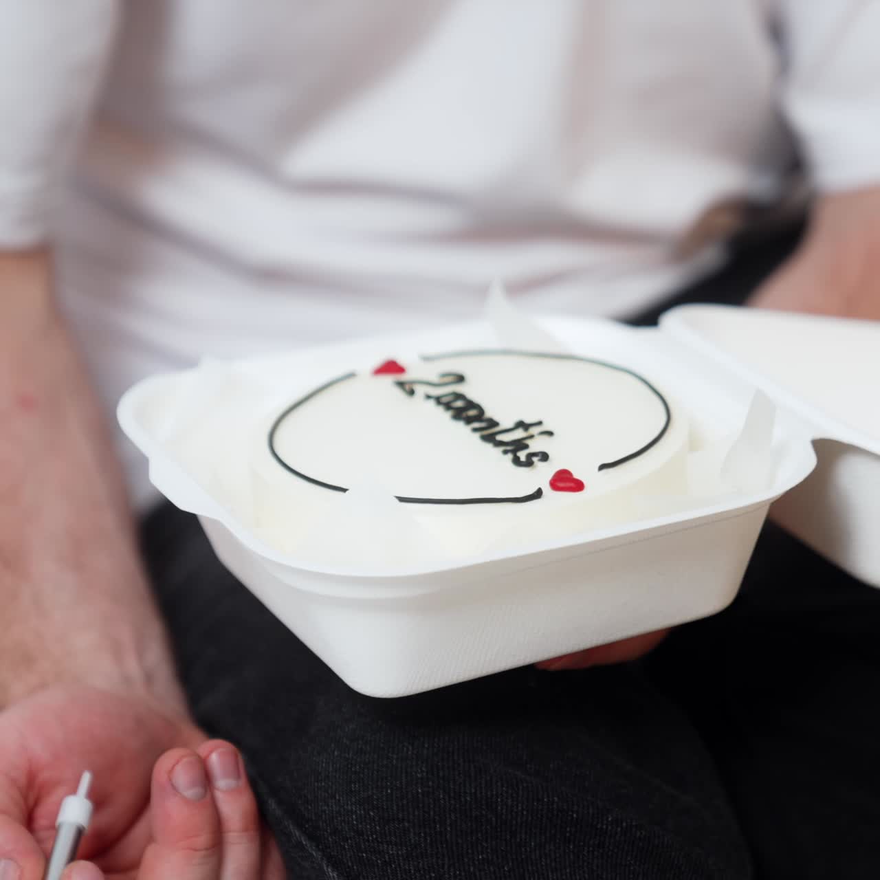 Unrecognized man holding a food box with a cake signed two months. Hand puts a candle into a cake. Close up