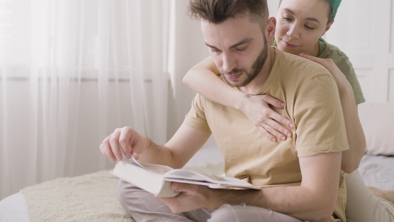 joven sentado en la cama y leyendo un libro mientras su novia lo acaricia suavemente por detrás