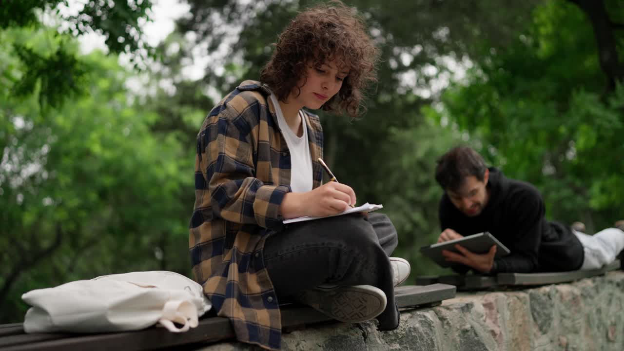 A brunette student girl with curly hair in a brown shirt sits on a bench and makes notes in a notebook near her fellow guy in the park