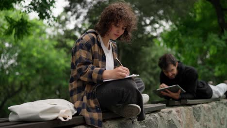 A-brunette-student-girl-with-curly-hair-in-a-brown-shirt-sits-on-a-bench-and-makes-notes-in-a-notebook-near-her-fellow-guy-in-the-park