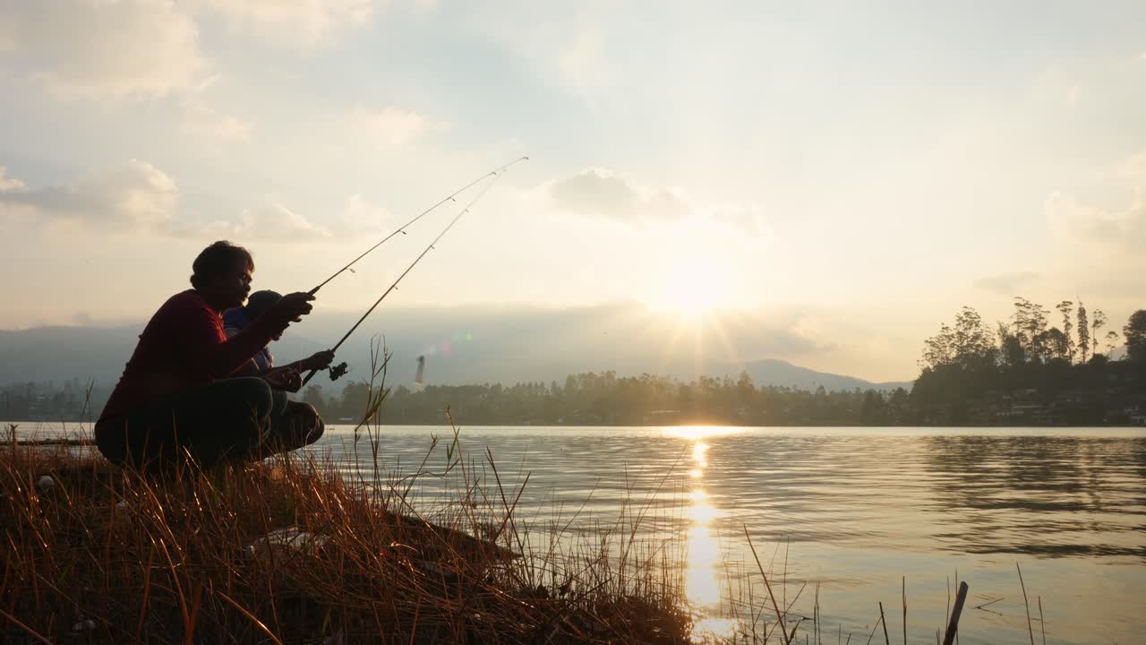 Two Men Crouch On The Lake Shore Throwing Fishing Rods Into The Lake
